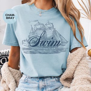 Pu&ograve; includere: T-shirt azzurra con un'illustrazione in stile vintage di una barca a vela e la parola "Swim" in corsivo. La maglietta ha maniche corte e scollo tondo.
