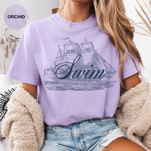 Pu&ograve; includere: T-shirt lilla chiaro con un'illustrazione vintage di una barca a vela e la parola "Swim" in corsivo. La maglietta &egrave; realizzata in un materiale morbido e confortevole.