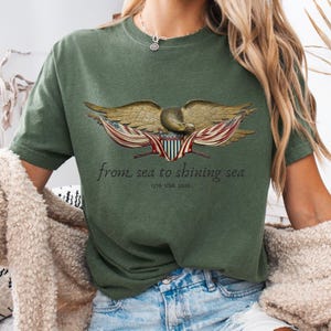 May include: Olive green t-shirt featuring a detailed illustration of an eagle with outstretched wings, incorporating elements of the American flag. The text "from sea to shining sea" and "1776 USA 2020" are printed below the eagle.