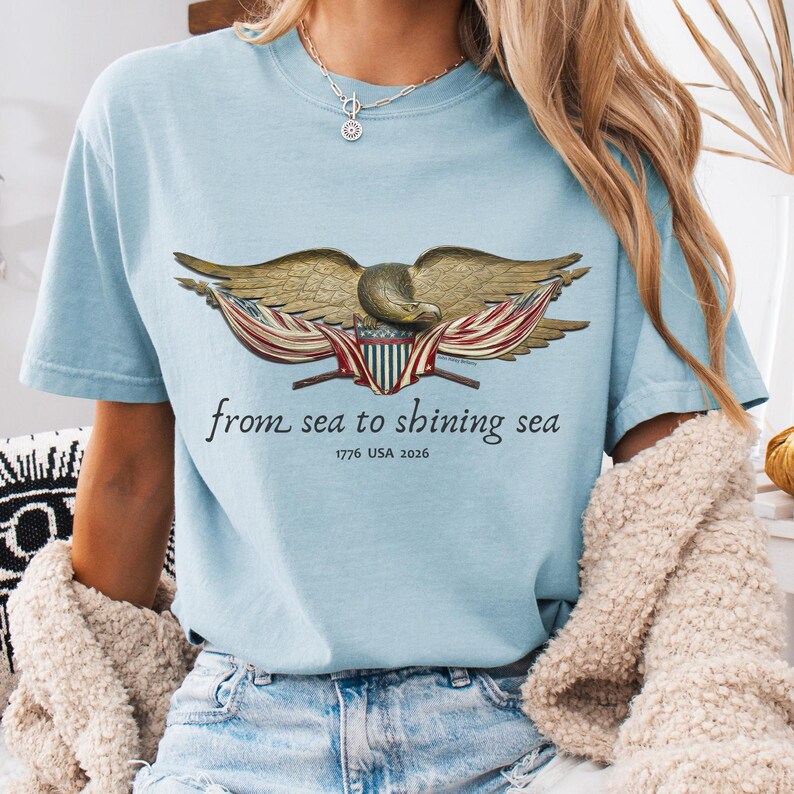 May include: Light blue t-shirt featuring a detailed illustration of an eagle with outstretched wings, holding a shield and flags. The text "from sea to shining sea" and "1776 USA 2026" are printed below the eagle. The shirt is made of soft material.