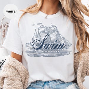 Pu&ograve; includere: T-shirt bianca con un'illustrazione blu di una barca a vela e la parola "Swim" in corsivo. La maglietta ha maniche corte e girocollo. La parola "WHITE" &egrave; in un cerchio bianco in alto a sinistra.