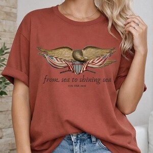 May include: Rust-colored t-shirt featuring a detailed eagle graphic with outstretched wings, a shield, and the text "from sea to shining sea" along with "1776 USA 2026". The design is centered on the front of the shirt.