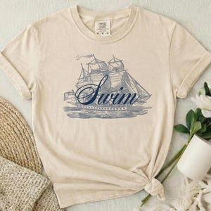 May include: Cream-colored t-shirt featuring a blue illustration of a sailboat with the word "Swim" in a cursive font. The shirt has short sleeves and a crew neck. The fabric appears soft and comfortable.