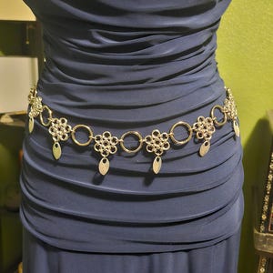 May include: A silver-toned metal belt with floral and oval links, and teardrop-shaped dangles. The belt is worn over a dark blue dress. The dress has a ruched design.