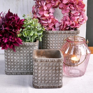 Könnte beinhalten: Arrangement dekorativer Artikel, darunter quadratische, gemusterte Pflanzgefäße mit Blumen und ein rosa Kranz. Eine rosa Glaslaterne mit einer brennenden Kerze befindet sich in der Nähe. Die Blumen sind tiefviolett und grün.