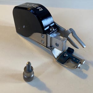 May include: A vintage Singer sewing machine attachment, featuring a black housing with the brand name in white lettering. The attachment is made of metal and has a silver finish, with a small, cylindrical screw next to it. This tool is likely used for sewing.