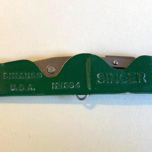May include: A vintage green Singer seam ripper with the text "SIMANCO U.S.A. 121834" and "SINGER" printed on the handle. The blade is silver and the tool has a small metal loop. The tool is closed and laying flat.