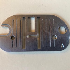 May include: A silver-colored sewing machine needle plate with a curved edge and multiple vertical slots. It has two small holes and a larger opening on one side. The letter "A" is stamped on the plate.