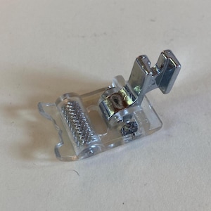 May include: A clear, plastic sewing machine foot with a silver metal attachment. The foot has a textured roller and is designed to guide fabric smoothly during sewing. Ideal for various sewing projects.