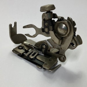 May include: A vintage, silver-toned sewing machine attachment. The metal piece has a complex design with multiple levers, a circular knob, and a spring mechanism. The attachment is designed to be mounted on a sewing machine.