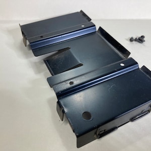 May include: Three black metal brackets with a rectangular shape and folded edges. Several small screws are next to the brackets. The brackets have multiple holes for mounting and are likely used for construction or repair.