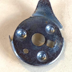 May include: A close-up of a silver-colored metal sewing machine bobbin case. The bobbin case has a circular shape with a central hole, a square opening, and three raised circular rivets. A curved tab extends from the top.