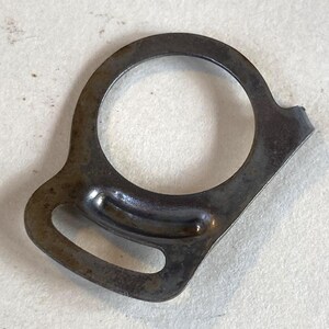 May include: A small, dark metal ring with a circular opening and a curved, oblong section. The metal has a worn, aged appearance with some discoloration. The ring has a small, protruding tab on one side.