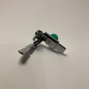 May include: A silver sewing machine foot with a green adjustment knob. The foot is designed for sewing a rolled hem.