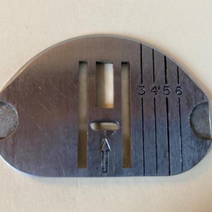 May include: A silver-colored sewing machine needle plate with a series of vertical lines and the numbers 3, 4, 5, and 6. The plate has a central rectangular opening and an arrow pointing upwards. Two semi-circular cutouts are on either side.