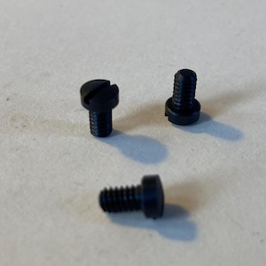 May include: Three small, black screws with slotted heads and threaded shafts. The screws are arranged on a light-colored surface, with one lying horizontally and the other two standing upright.