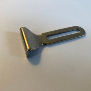 May include: A small, silver-colored metal buckle with a rectangular slot and a curved, angled end. The buckle is designed for use with straps or belts and is likely used in clothing or accessories.