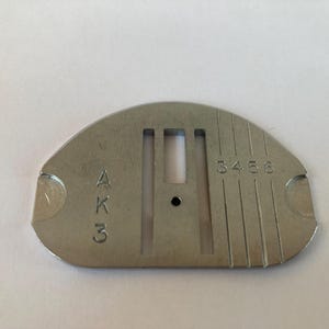 May include: A silver-colored sewing machine needle plate with the markings "AK3" and "3456". The plate has a curved top and a series of slots and a small hole in the center. The plate is made of metal.