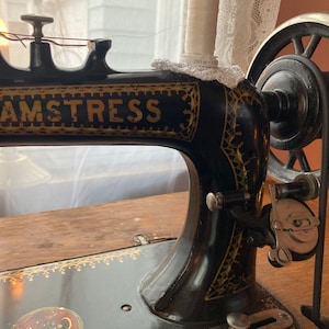 May include: A vintage black sewing machine with gold accents. The machine has a large wheel on the right side and the word "SEAMSTRESS" printed on the front.