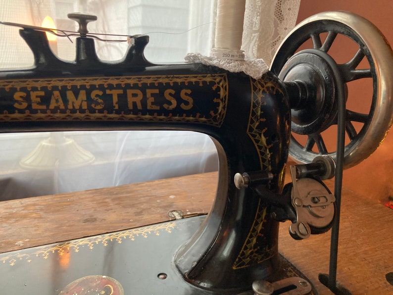 May include: A vintage black sewing machine with gold accents. The machine has a large wheel on the right side and the word "SEAMSTRESS" printed on the front.