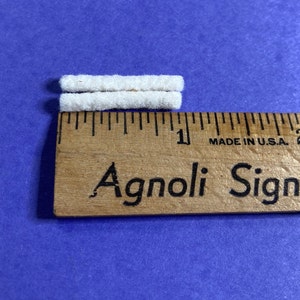 May include: Two white cotton cord ends, each about 1 inch long, are placed next to a wooden ruler with the text "Agnoli Sign" printed on it.