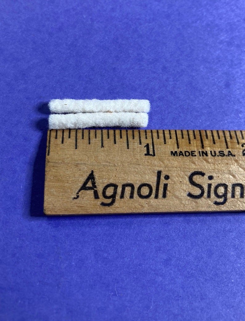 May include: Two white cotton cord ends, each about 1 inch long, are placed next to a wooden ruler with the text "Agnoli Sign" printed on it.