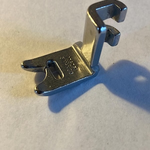 May include: A silver-colored metal sewing machine presser foot with a two-pronged front and a vertical attachment shank. The metal surface features the stamped text 105151 SIMANKO, indicating the specific model and brand of this sewing component.