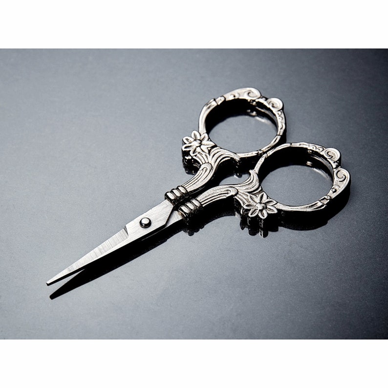 May include: Silver-toned decorative scissors with ornate floral details. The scissors have a pointed tip and a small round knob in the center.