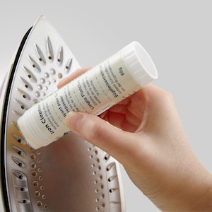 May include: A white tube of iron cleaner with the text "Iron Cleaner", "Repasser", "Limpia Planchas", "Bügeleisenreiniger", and "60g" on the label. The tube is being used to clean the soleplate of an iron.