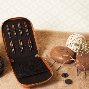 May include: An open brown leather case with a zip closure, containing a set of wooden knitting needles. The case is lying on a beige fluffy surface with two brown stone-like objects and two black circular objects.