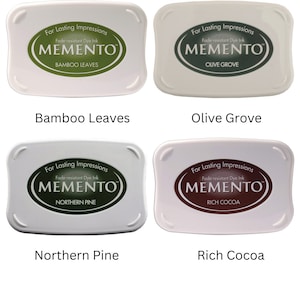 Pode incluir: Quatro almofadas de tinta para carimbar, cada uma com uma cor e um nome diferentes. As cores s&atilde;o verde, verde escuro, marrom e marrom claro. Os nomes s&atilde;o Folhas de Bambu, Olival, Pinho do Norte e Cacau Rico. Cada almofada tem o texto "Para impress&otilde;es duradouras" e "Tinta de corante resistente ao desbotamento" e o logotipo "MEMENTO".