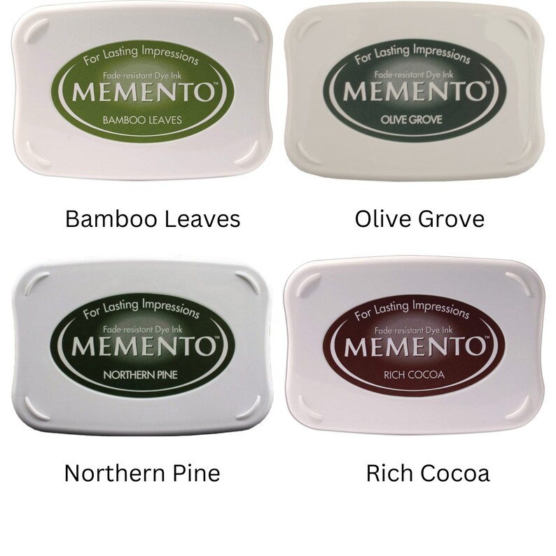 Pode incluir: Quatro almofadas de tinta para carimbar, cada uma com uma cor e um nome diferentes. As cores s&atilde;o verde, verde escuro, marrom e marrom claro. Os nomes s&atilde;o Folhas de Bambu, Olival, Pinho do Norte e Cacau Rico. Cada almofada tem o texto "Para impress&otilde;es duradouras" e "Tinta de corante resistente ao desbotamento" e o logotipo "MEMENTO".