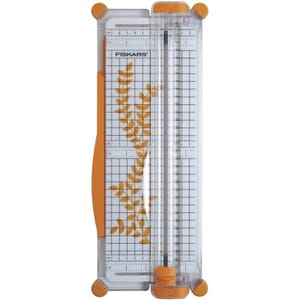 May include: A clear and orange Fiskars paper trimmer with a grid and ruler markings. The trimmer has a sliding blade and a decorative leaf design. The blade is designed to cut paper up to 12 inches.