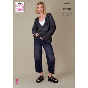 Può includere: Una donna indossa un maglione Aran grigio scuro con scollo a V sopra un top bianco, abbinato a jeans scuri e sandali neri. Le misure del maglione sono 81-107 cm. L'immagine include il logo King Cole e il numero 6446.