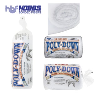 Op de afbeelding: Een witte rol Hobbs Poly-Down Premium Polyester quiltvulling. De vulling is gemakkelijk te quilten, heeft een donsachtige zachtheid, is machinewasbaar en hypoallergeen. Het etiket zegt "Hobbs Poly-Down Premium Polyester" en "Gemakkelijk te quilten".