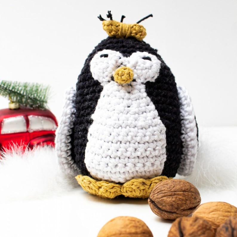 May include: A crocheted penguin toy with black and white fur, a yellow beak, and a yellow crown. The penguin is sitting on a white surface with walnuts.
