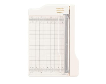 May include: A white paper trimmer with a grid and ruler markings. The trimmer has a silver blade guide and the text "vaessen creative" in gold. The grid is marked in inches and centimeters.