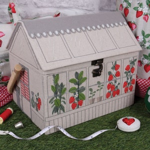May include: A fabric sewing box shaped like a greenhouse, with a gray roof and strawberry illustrations. Other sewing accessories include a pin cushion, tape measure, scissors, and thread. A matching tote bag and knitting needle holder are also visible.