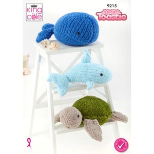 Peut inclure: Trois animaux en peluche tricotés : une baleine bleue, un requin bleu clair et une tortue verte et beige. Les jouets sont exposés sur un tabouret blanc. Le texte "Jumbo Toastie" est visible.