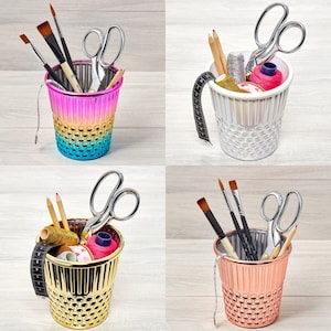 Könnte beinhalten: Vier farbige, metallene Behälter in Form von Fingerhüten mit verschiedenen Nähutensilien, darunter Scheren, Nadeln, Garn und Bleistifte. Die Behälter sind mit einem strukturierten Muster verziert und haben einen kontrastierenden Farbrand.