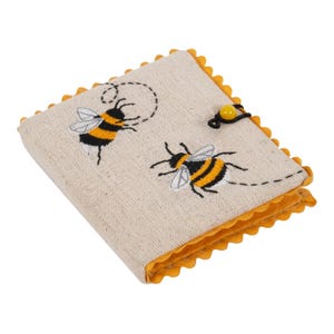 May include: A beige fabric notebook featuring embroidered yellow and black bees. The notebook has a yellow scalloped edge and a yellow button closure. The cover displays two embroidered bees with dotted flight paths.