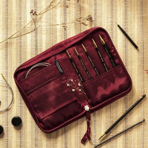 May include: A burgundy zippered case with a set of six black and gold knitting needles inside. The case has a label that reads "Lantern Moon Handmade".