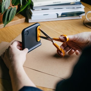 May include: A black and orange paper trimmer with scissors cutting a piece of brown paper. The trimmer has a rectangular shape with a curved top and a yellow guide. The scissors are orange and silver.