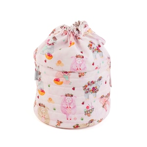 May include: A pink quilted yarn storage bag with a drawstring closure. The bag is covered in a repeating pattern of sheep wearing floral crowns and holding knitting needles. The bag has a small tag that says "hobby gift".