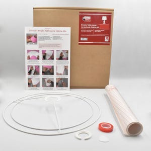 May include: A lampshade making kit, featuring a brown cardboard box, instructions, and a white wire frame. The kit includes a roll of clear adhesive film, red tape, and other components for crafting a lampshade.