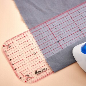 May include: A clear plastic ruler with red markings for measuring fabric. The ruler has a curved edge and is labeled "Hemline" with the website address. The ruler is placed on top of a piece of gray fabric.