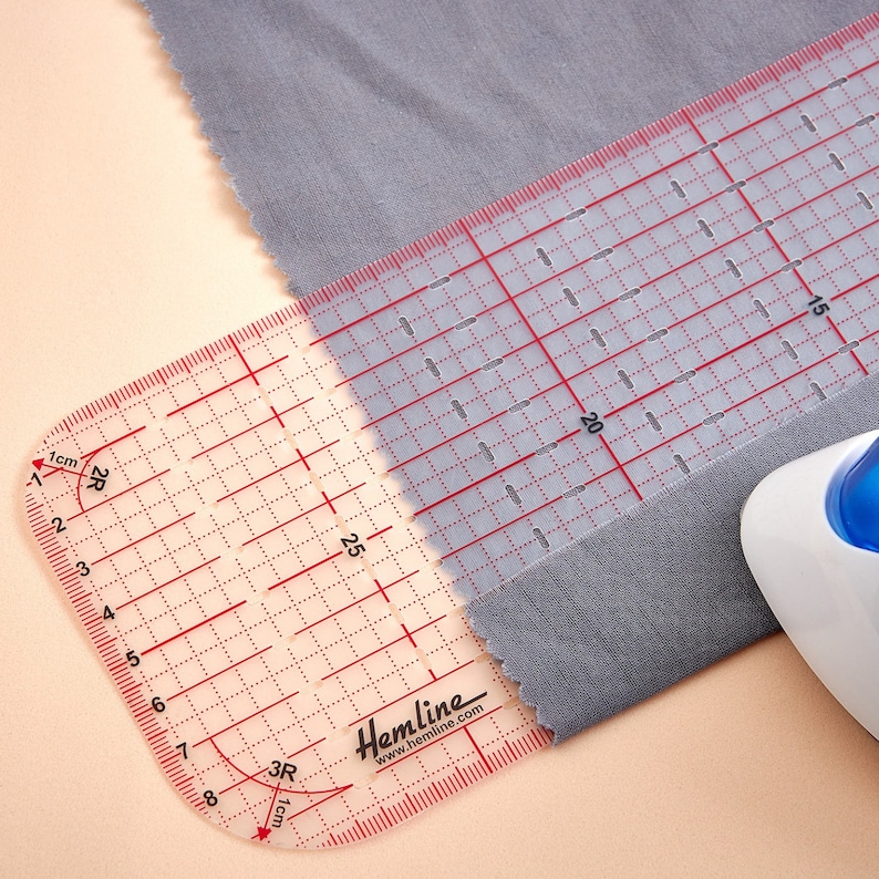 May include: A clear plastic ruler with red markings for measuring fabric. The ruler has a curved edge and is labeled "Hemline" with the website address. The ruler is placed on top of a piece of gray fabric.