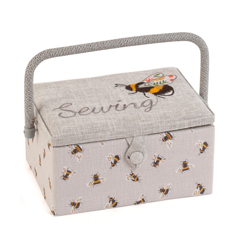 Puede incluir: Una caja de costura de tela gris con asa. La caja tiene un patr&oacute;n de abejas y flores y la palabra "Costura" bordada en la tapa.