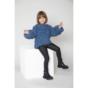 May include: A young person wearing a blue fuzzy sweater and black pants sits on a white cube. They are wearing black boots with buckles.