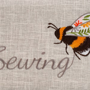 Puede incluir: Una tela de lino beige con la palabra "Costura" bordada en hilo gris. Una abeja bordada estilizada con rayas negras, amarillas y blancas vuela sobre la palabra. La abeja tiene alas florales con acentos rosas, rojos, amarillos y verdes.